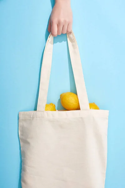 Vista recortada de la mujer sosteniendo limones amarillos maduros en bolsa de algodón sobre fondo azul - foto de stock