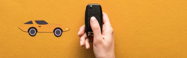 Cropped view of woman holding car key near paper cut auto on orange background with copy space, panoramic shot — Stock Photo
