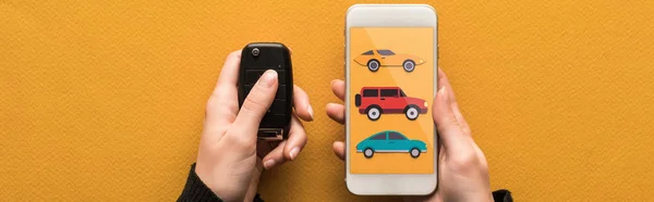 Cropped view of woman holding car key and smartphone with car rent app on screen on orange background, panoramic shot — Stock Photo