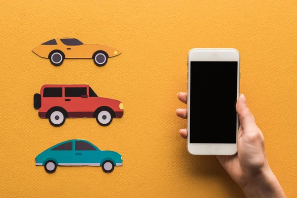 Cropped view of woman holding smartphone with blank screen near paper cut cars, location mark on orange background — Stock Photo