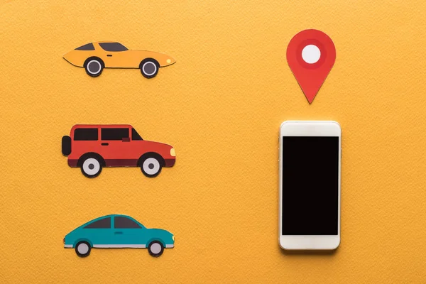 Вид сверху на разрезанные бумагой автомобили, метка местоположения рядом со смартфоном с пустым экраном на оранжевом фоне — стоковое фото