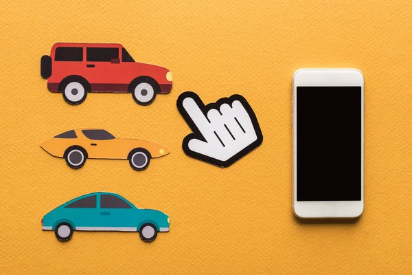 Vista superior de coches cortados en papel, apuntando con la mano y el teléfono inteligente sobre fondo naranja - foto de stock