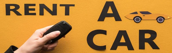 Vista cortada de mulher segurando chave perto de papel cortado alugar um carro lettering no fundo laranja, tiro panorâmico — Fotografia de Stock
