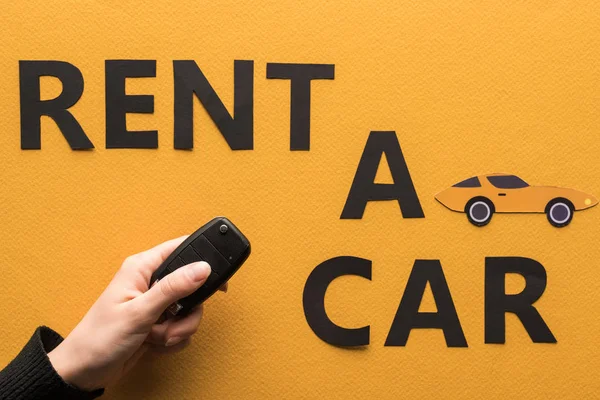 Corte vista de mulher segurando chave perto de papel cortar alugar um carro lettering no fundo laranja — Fotografia de Stock