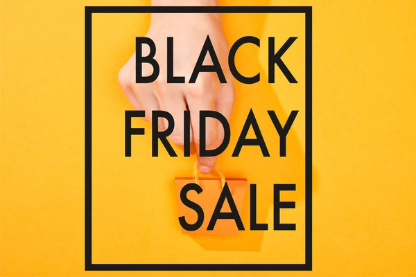 Corte vista de mão segurando pequeno saco de compras no fundo laranja brilhante com preto sexta-feira venda ilustração — Fotografia de Stock