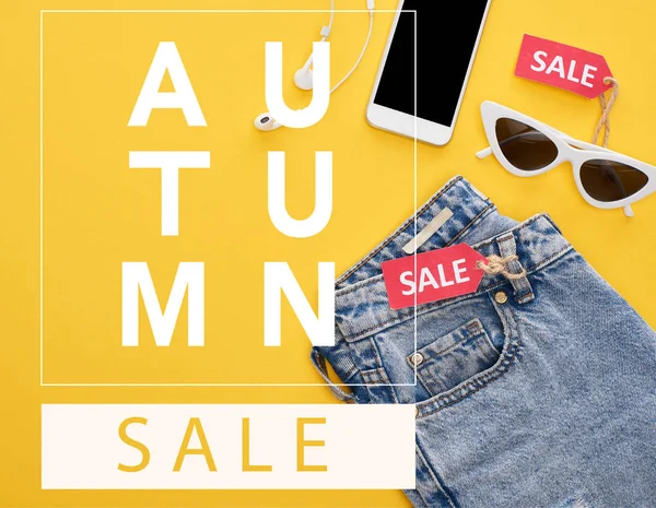 Vista superior de jeans, gafas y smartphone con auriculares con etiquetas de venta sobre fondo amarillo con ilustración de venta de otoño - foto de stock