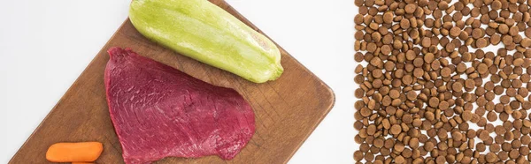 Vista superior de alimentos secos para mascotas cerca de carne cruda, zanahoria y calabacín en una tabla de cortar de madera aislada en blanco, plano panorámico - foto de stock