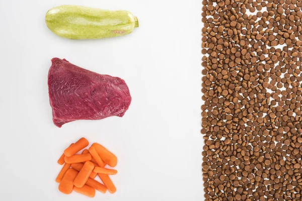 Tendido plano con comida seca para mascotas cerca de carne cruda, zanahoria y calabacín aislados en blanco - foto de stock