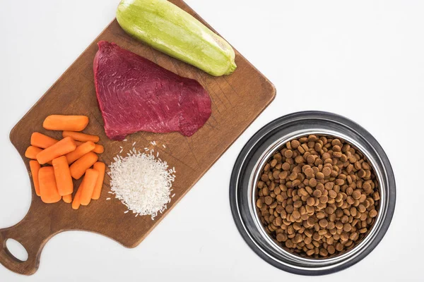Vista dall'alto di cibo secco per animali domestici e carne cruda, riso, carote e zucchine su tagliere in legno isolato su bianco — Foto stock