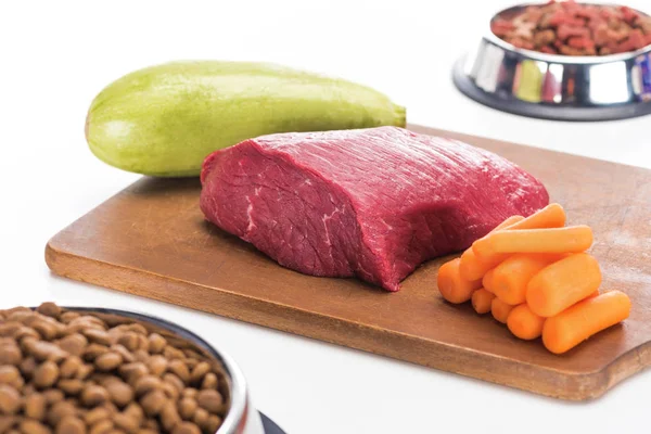 Selective focus of dry pet food in bowls near raw meat, carrot and zucchini on wooden cutting board isolated on white — Stock Photo