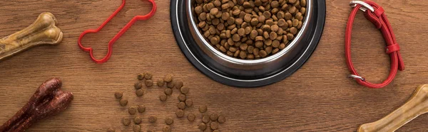 Top view of dry pet food in silver bowl near bones, collar and pet toy on wooden table, panoramic shot — Stock Photo