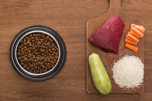 Draufsicht auf Trockenfutter in silberner Schüssel in der Nähe von rohem Fleisch, Zucchini, Reis und Karotten auf Holztisch — Stockfoto