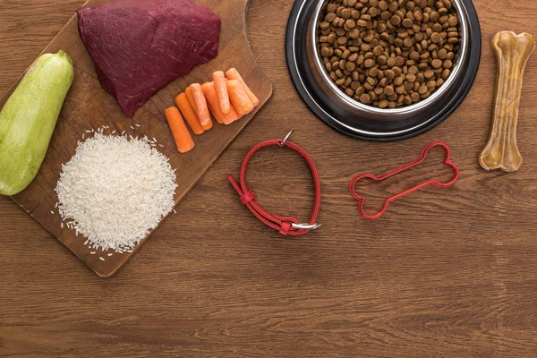 Top view of dry pet food in silver bowl near raw meat, carrot, rice, zucchini, bone, collar and pet toy on wooden table — Stock Photo