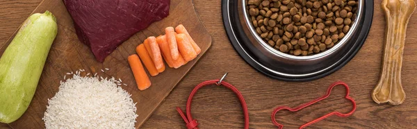 Top view of dry pet food in silver bowl near raw meat, carrot, rice, zucchini, bone, collar and pet toy on wooden table, panoramic shot — Stock Photo