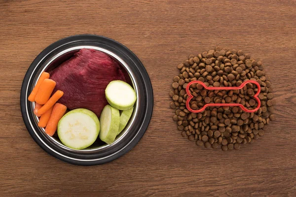 Vista superior de alimentos secos para mascotas con hueso cerca de verduras crudas y carne en un tazón sobre una mesa de madera - foto de stock