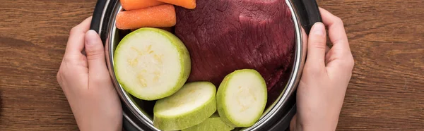 Cropped view of woman holding pet bowl with raw meat and vegetables on wooden background, panoramic shot — Stock Photo