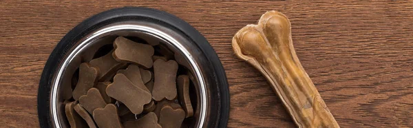 Top view of dry pet food in silver bowl near bone on wooden table, panoramic shot — Stock Photo