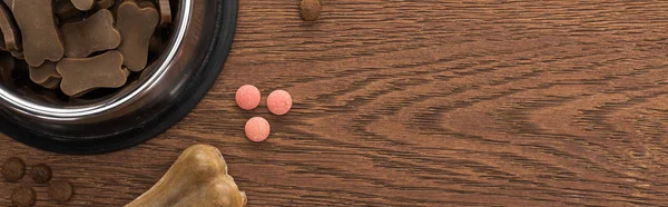 Vista superior de comida seca para mascotas en tazón de plata cerca del hueso y vitaminas rosadas en la mesa de madera, plano panorámico - foto de stock