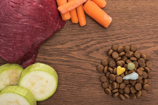 Top view of raw meet, zucchini and carrot near vitamins on dry pet food on wooden table — Stock Photo