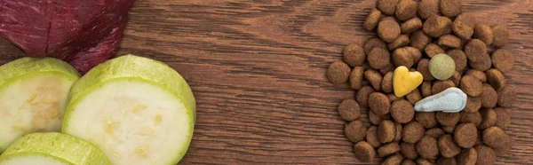 Top view of raw meet, zucchini near vitamins on dry pet food on wooden table, panoramic shot — Stock Photo
