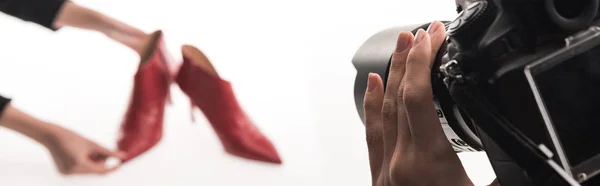 Обрезанный вид коммерческих фотографов, делающих коммерческую фотосессию женской обуви на красных каблуках на белом, панорамный снимок — стоковое фото