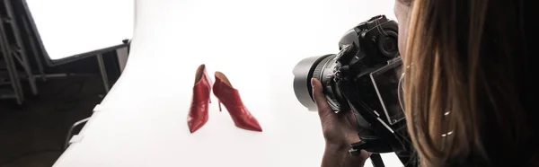 Vista recortada del fotógrafo haciendo sesión de fotos comerciales de zapatos de tacón rojo femenino en blanco, tiro panorámico - foto de stock