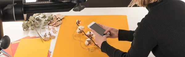 Cropped view of photographer taking photo of composition with cotton flower and accessories on smartphone, panoramic shot — Stock Photo