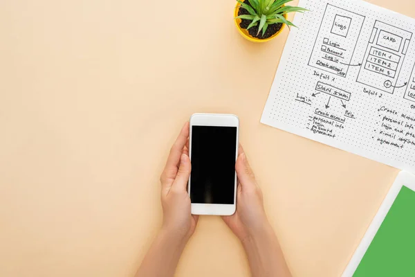 Abgeschnittene Ansicht einer Frau mit Smartphone in der Nähe von Website-Design-Vorlage, digitalem Tablet und grüner Pflanze auf beigem Hintergrund — Stockfoto