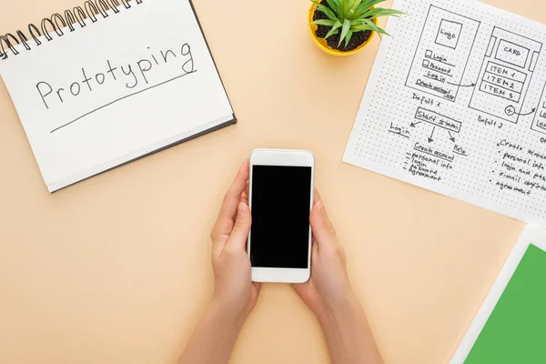 Cropped view of woman holding smartphone near notebook with prototyping lettering, website design template, digital tablet and green plant on beige background — Stock Photo