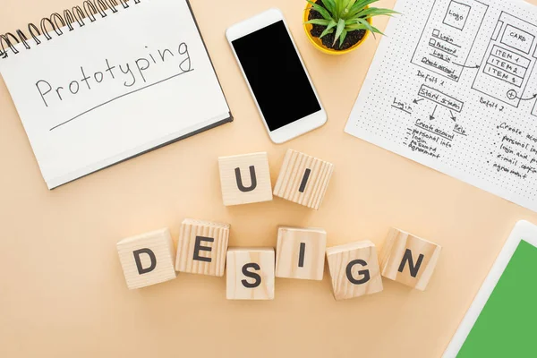 Vista dall'alto di gadget vicino a blocchi di legno con ui lettering design, modello di design del sito web, notebook con scritte di prototipazione e pianta verde su sfondo beige — Foto stock