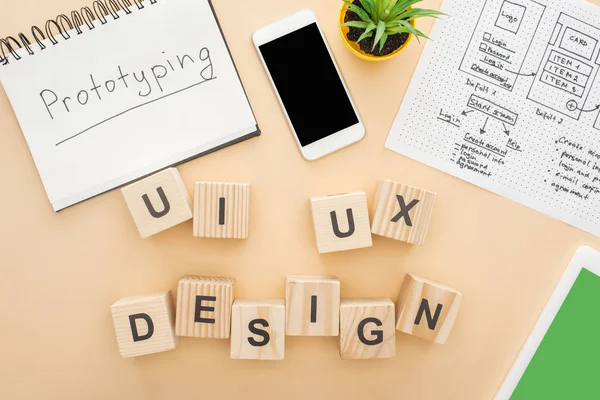 Vista superior de gadgets perto de blocos de madeira com letras de design ui e ux, modelo de design de site, notebook com prototipagem lettering e planta verde no fundo bege — Fotografia de Stock