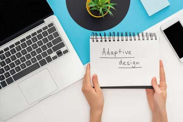 Cropped view of woman holding notebook with adaptive design lettering near laptop, smartphone, plant on abstract geometric background — Stock Photo