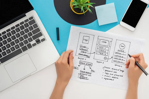 Vista recortada de la mujer que sostiene la plantilla de diseño del sitio web y la pluma de fieltro cerca de la computadora portátil, teléfono inteligente, planta sobre fondo geométrico abstracto - foto de stock