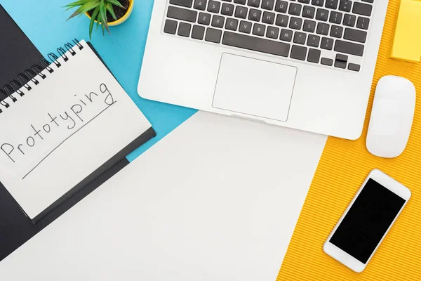 Top view of notepad with prototyping lettering, laptop, computer mouse, smartphone, plant on abstract geometric background — Stock Photo