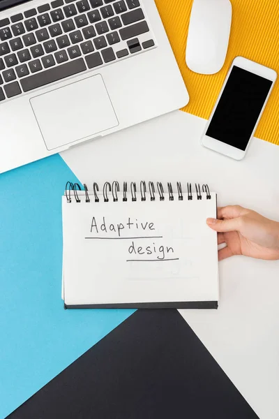 Cropped view of woman holding notebook with adaptive design lettering near laptop, smartphone, computer mouse on abstract geometric background — Stock Photo