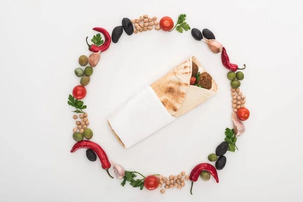 Top view of chickpea, garlic, cherry tomatoes, parsley, olives, chili pepper, green onion arranged in round frame around falafel in pita on white background — Stock Photo