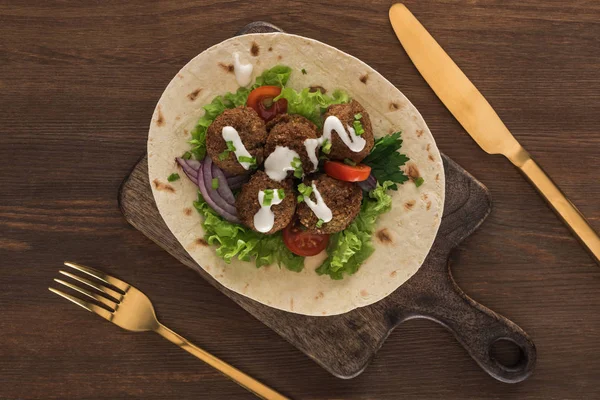 Top view of falafel with vegetables and sauce on pita near cutlery on wooden table — Stock Photo
