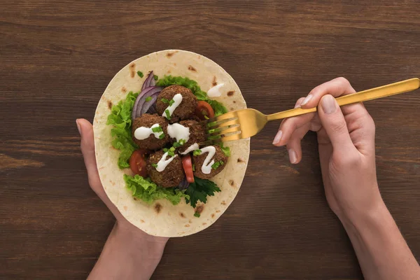 Vista ritagliata della donna che mangia falafel con verdure e salsa sulla pita con forchetta sul tavolo di legno — Foto stock