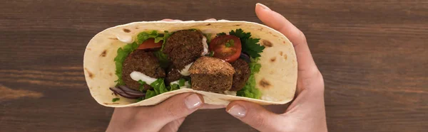 Vista recortada de la mujer sosteniendo falafel con verduras y salsa en pita sobre mesa de madera, plano panorámico - foto de stock