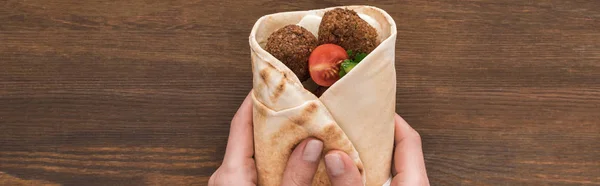 Vista recortada de la mujer sosteniendo falafel con verduras y salsa en pita sobre mesa de madera, plano panorámico - foto de stock