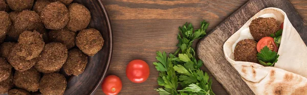 Top view of falafel with vegetables and sauce in pita near balls, parsley, tomatoes on wooden table, panoramic shot — Stock Photo