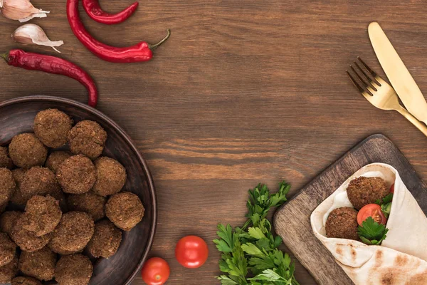 Top view of falafel with vegetables and sauce in pita near balls, parsley, tomatoes, chili pepper, garlic and cutlery on wooden table — Stock Photo