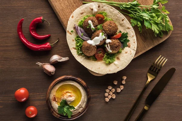 Vista superior de falafel com legumes em pita perto de talheres, especiarias e húmus em mesa de madeira perto de especiarias — Fotografia de Stock