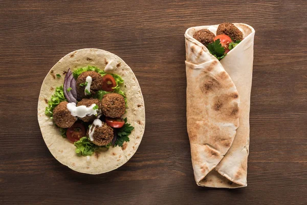 Vista superior de falafel con verduras y salsa envueltas y desenvueltas en pita sobre mesa de madera - foto de stock