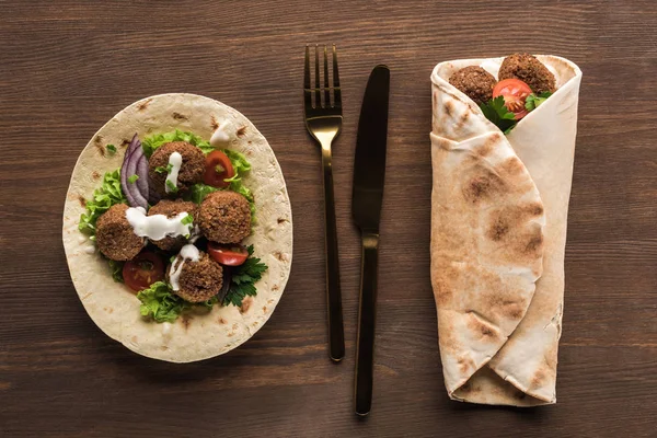 Top view of falafel with vegetables and sauce wrapped and unwrapped in pita on wooden table with cutlery — Stock Photo