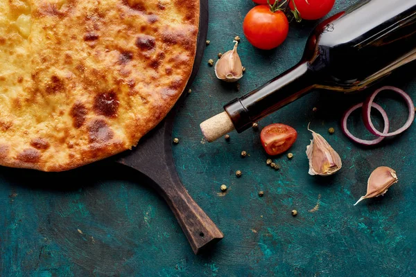 Imereti khachapuri on chopping board with wine bottle and vegetables on textured green background — Stock Photo