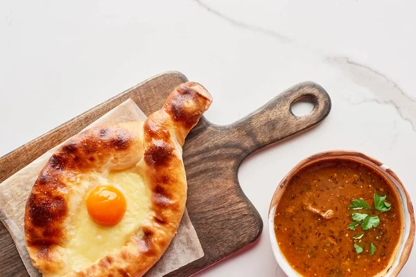 Top view of adjarian khachapuri on cutting board and soup kharcho on marble texture — Stock Photo