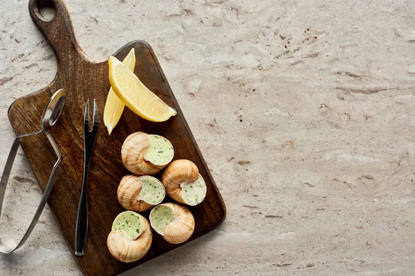 Vista superior de deliciosas escargotas cocidas servidas en tabla de cortar de madera con rodajas de limón y pinzas sobre fondo de piedra - foto de stock