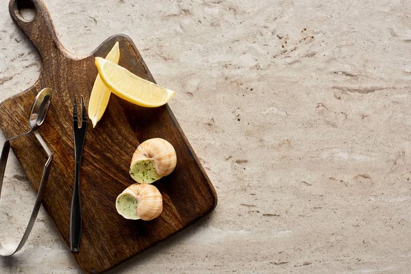 Top view of delicious cooked escargots served on wooden cutting board with lemon slices and tweezers on stone background — Stock Photo