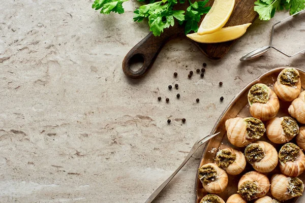 Vista superior de deliciosas escargotas cozidas com fatias de limão, salsa, pimenta preta e pinças no fundo de pedra — Fotografia de Stock